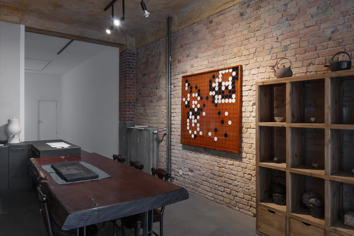 Installationsansicht: Raum mit Backsteinwand, Holztisch, Regalen und einem Go-Brett an der Wand