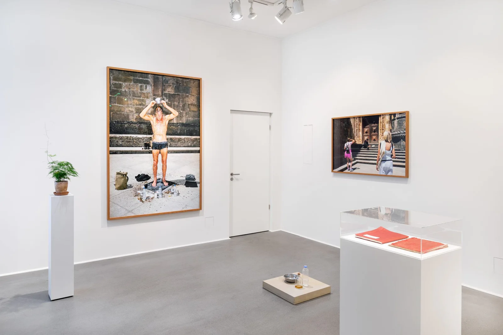 Installation view: white room with the photograph of a man in swimming trunks, another framed photograph and a display case with red books