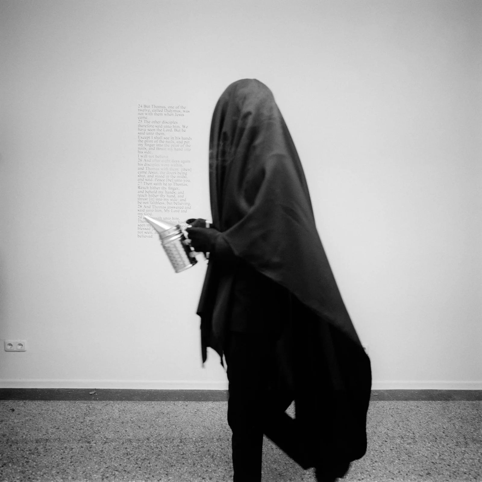 Max Schwarzmann: a figure veiled in black holding a book in front of a white wall with a text panel, black and white