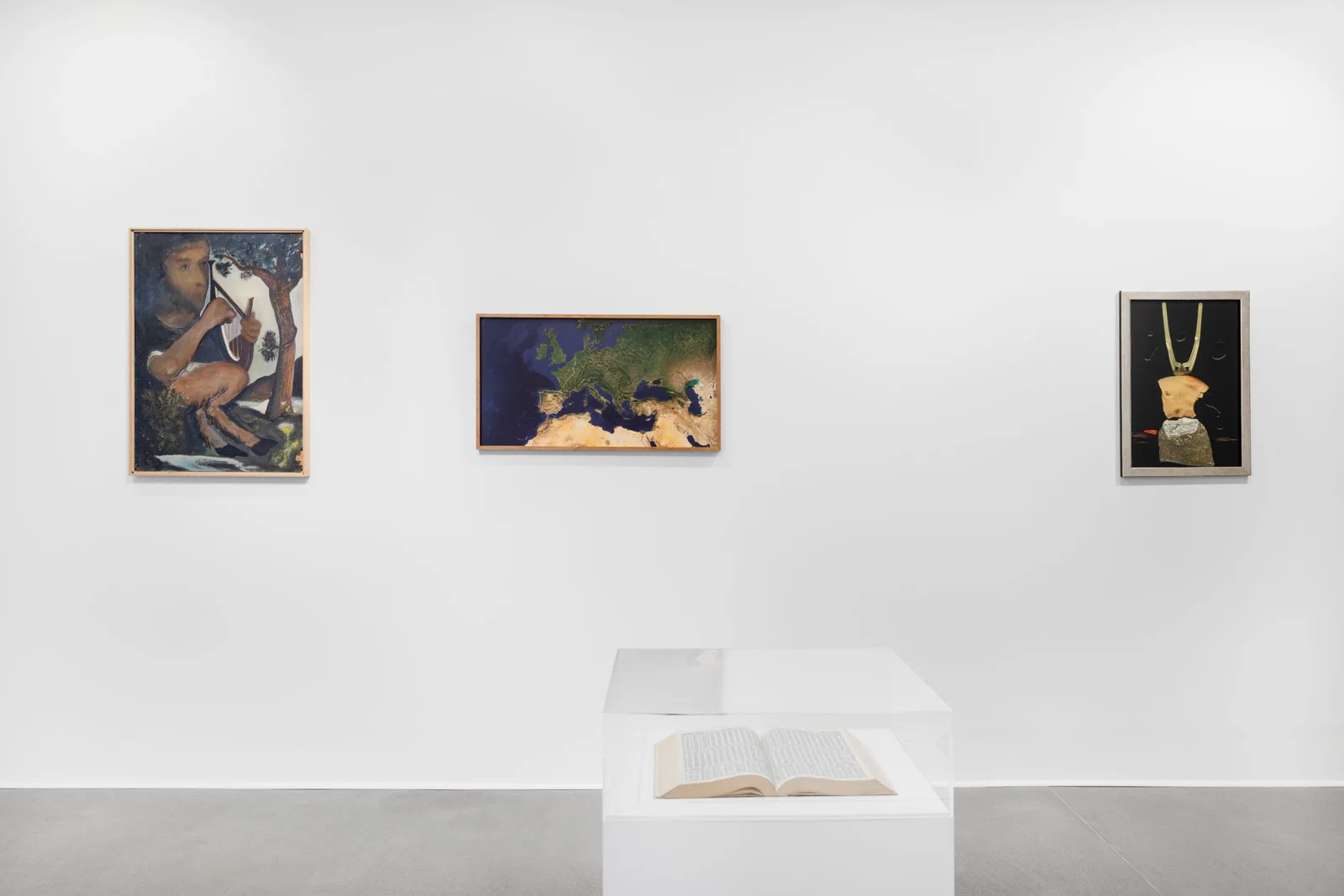 Exhibition view: three paintings by Hilarion Manero on a white wall, in front of them an open book on a plinth