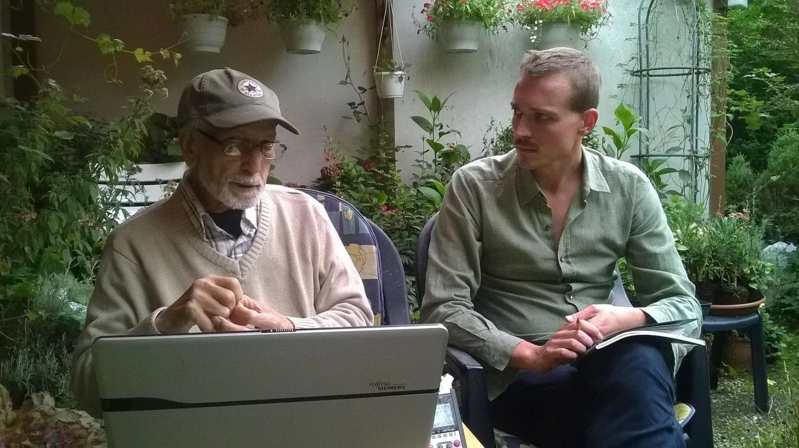Julian Malte Schindele im Interview mit El Arbi Bouqdib an einem Laptop in einem Hof voller Pflanzen