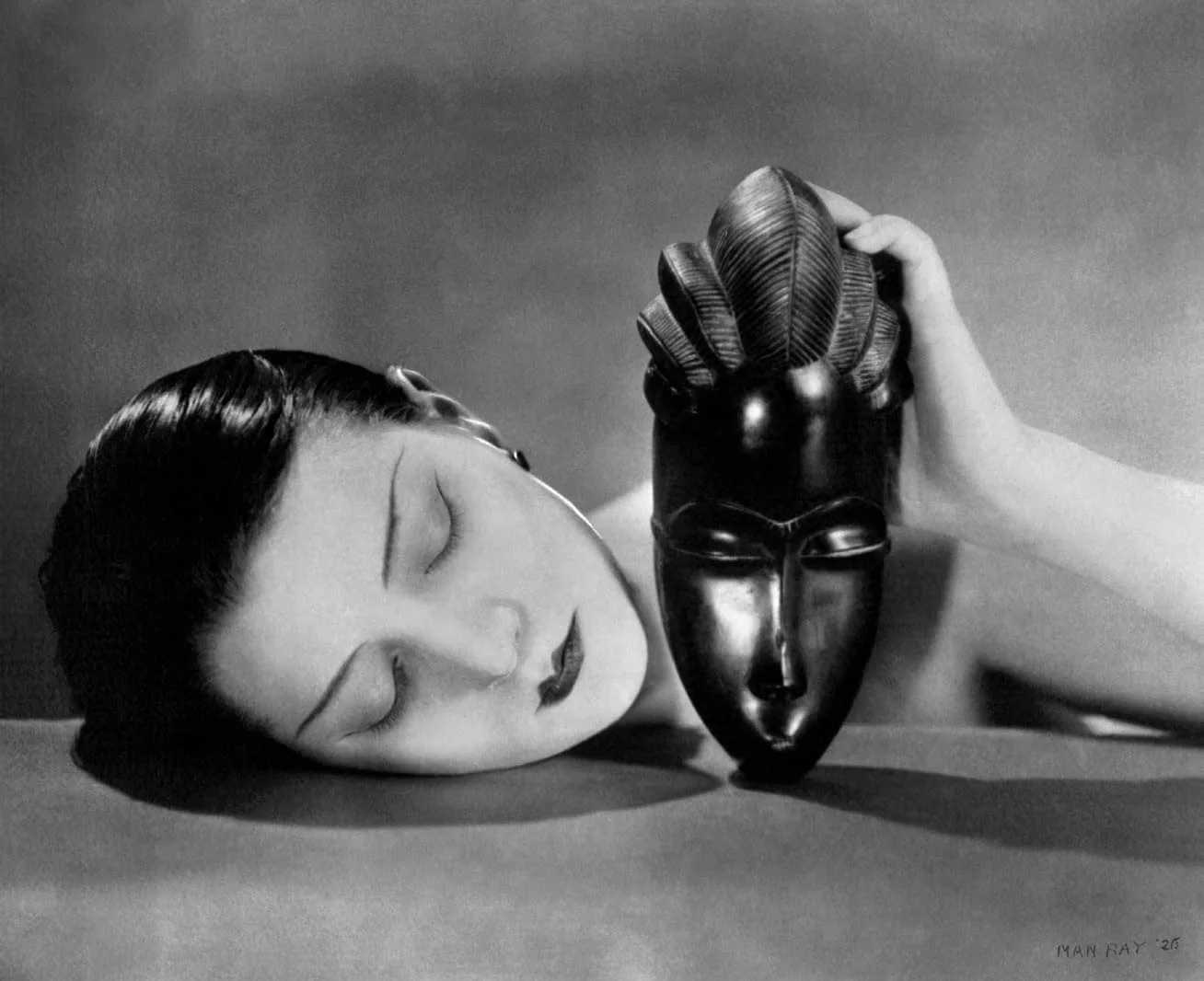 Man Ray, Noire et Blanche, 1926: black and white photograph of a woman resting her head on a table and holding an African mask
