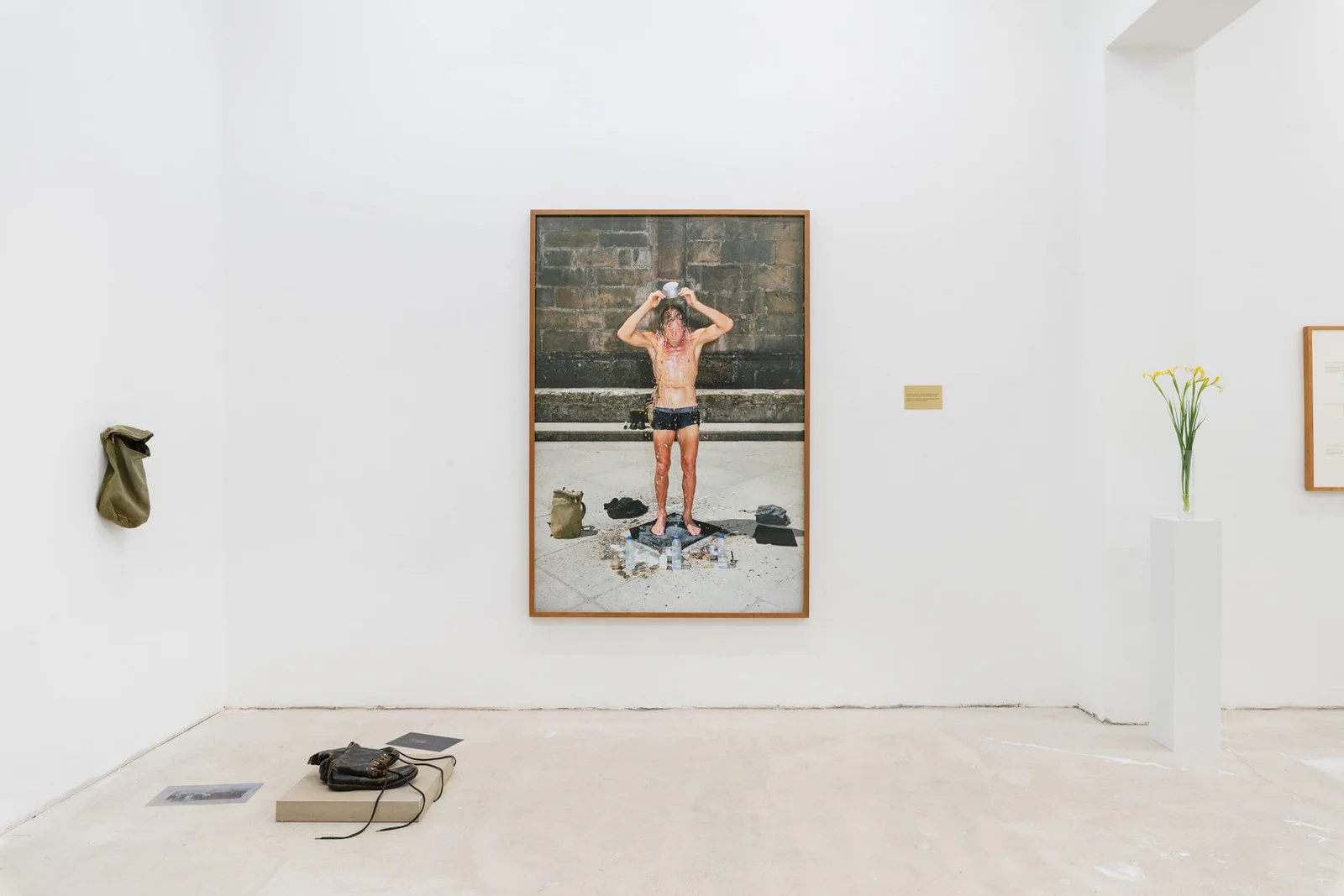 Die Dritte Waschung oder die Honigtaufe (Marianische Antiphonen I): gerahmte Fotografie eines Mannes in Badehose vor einer Mauer, daneben Messingtafel und Blumen