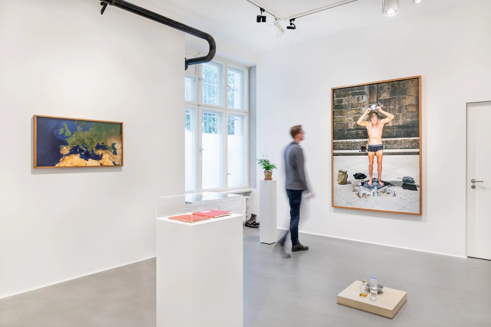 Installation view: a visitor in a white room with a satellite map of Europe, photographs and a display case with red books
