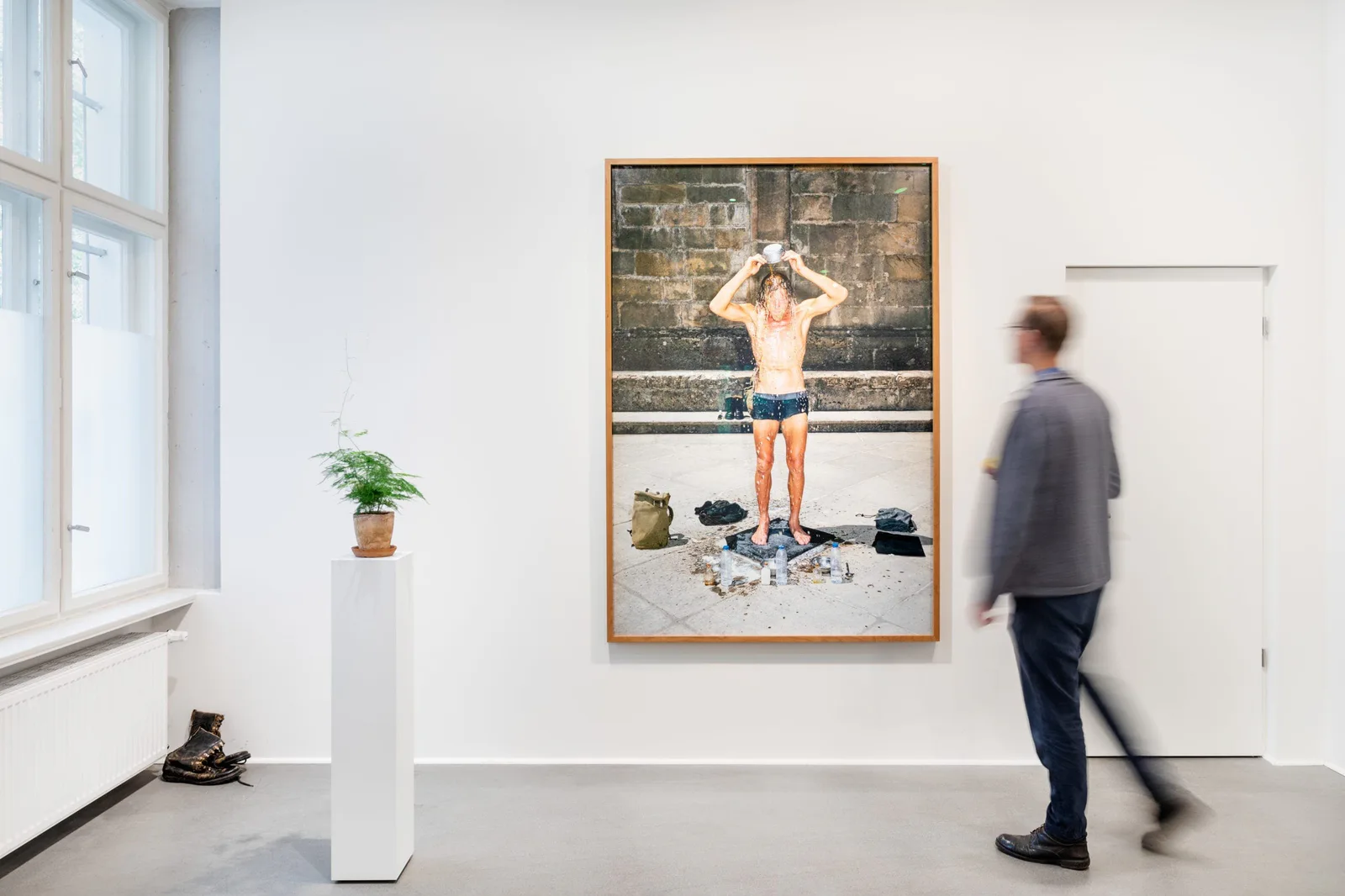 Installation view: a visitor in front of the photograph of a man in swimming trunks, a pair of boots on the floor in front