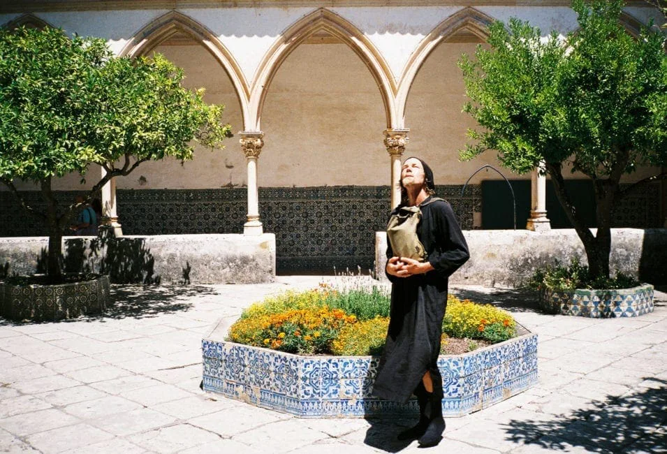 GABRIEL: a man in black standing in an arcaded courtyard next to a flower bed, looking upwards